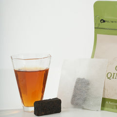Glass of amber liquid next to a tea bag and black tea brick on a white surface.