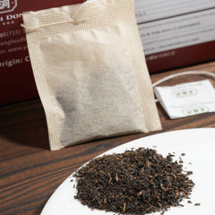 Tea bag and loose tea on a white plate with a wooden surface and product packaging in the background.